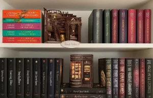 Étagère de bibliothèque décorative avec des romans de Sarah J. Maas et des serre-livres lumineux miniatures de type "book nook" créant une ambiance magique.