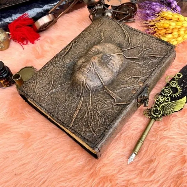 Grimoire artisanal en cuir marron avec un visage humain sculpté en relief sur la couverture, posé sur une fourrure rose à côté d'un stylo plume steampunk doré.