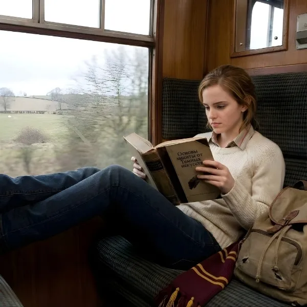 Hermione Granger assise dans le Poudlard Express, lisant attentivement un livre ancien sur la défense contre les forces du Mal, avec son sac à dos et son écharpe Gryffondor à ses côtés.