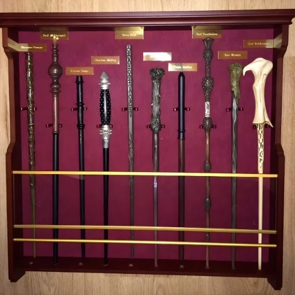 Présentoir mural en bois contenant une collection de dix baguettes magiques de différentes longueurs sur fond de velours bordeaux. Chaque baguette est accompagnée d'une plaque nominative dorée, incluant celles de Dumbledore et Voldemort, protégées par des barres de sécurité dorées.