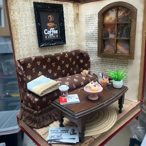 Une scène de salon miniature installée à l'intérieur d'un livre ouvert, comprenant un canapé floral marron, une table basse en bois avec des pâtisseries factices, un petit cadre "Coffee House" et une fenêtre ouvrant sur un paysage miniature.