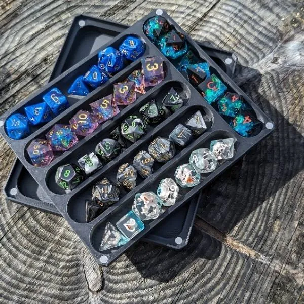 Un plateau de rangement noir compartimenté contenant plusieurs sets de dés de jeu de rôle variés (bleus, galaxie, transparents) posé sur une table en bois rustique.