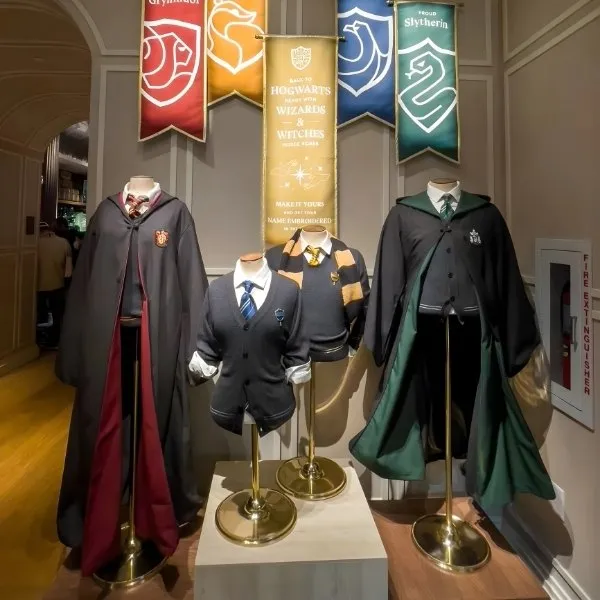 Exposition de quatre uniformes scolaires de Poudlard sur mannequins, incluant les robes et cardigans des maisons Gryffondor, Serpentard, Serdaigle et Poufsouffle, surmontés de leurs bannières respectives dans une boutique officielle Harry Potter.