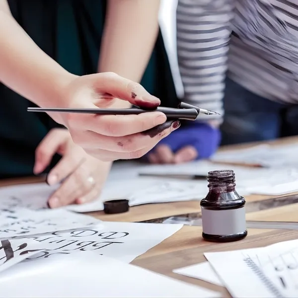 Une personne tenant un porte-plume au-dessus d'un flacon d'encre lors d'un atelier de calligraphie artistique sur papier.