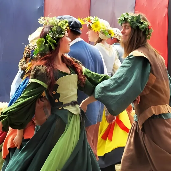 Groupe de jeunes gens en costumes médiévaux dansant lors d'une fête champêtre, portant des couronnes de fleurs et de feuillage, avec des robes vertes et des tuniques artisanales.