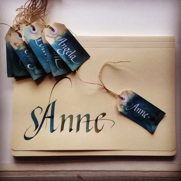 Étiquettes de cadeaux artisanales avec calligraphie élégante à l'encre blanche sur fond dégradé bleu et kraft, accompagnées d'une grande enveloppe portant le prénom "Anne" écrit à la main
