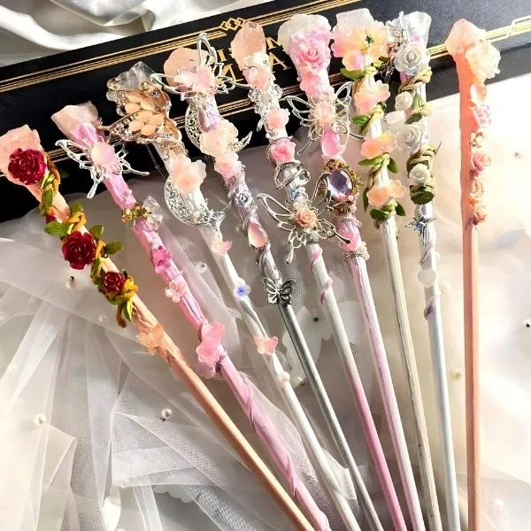 Collection de baguettes magiques artisanales ornées de fleurs roses, cristaux de quartz et papillons argentés sur fond de voile blanc